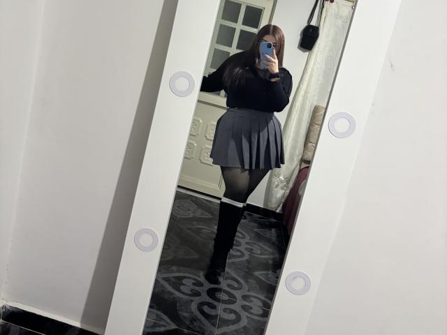 VenusxMusa mit langen, braunen Haaren, einem schwarzen, langärmligen Hemd, einem grauen, gefalteten Rock, schwarzen Strumpfhosen und schwarzen, kniehohen Stiefeln, macht ein Selfie vor einem Spiegel und zeigt ihren Hintern..