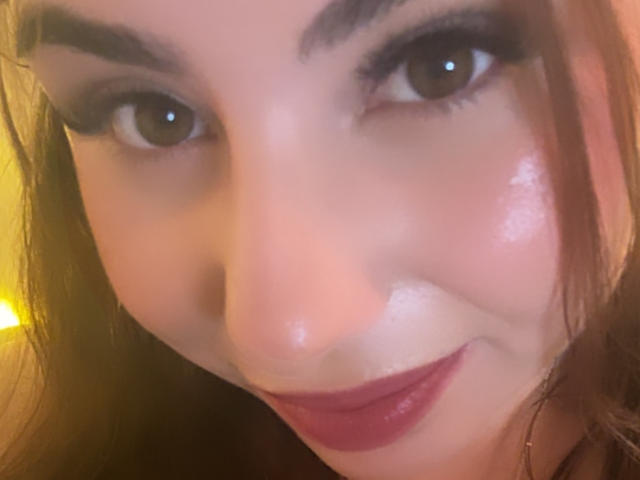 Das Gesicht von VenusxMusa mit glänzenden braunen Augen, dicken Augenbrauen und strahlender Haut. Sie trägt dunkelroten Lippenstift und hat ein leichtes Lächeln. Ihr braunes Haar ist teilweise sichtbar..