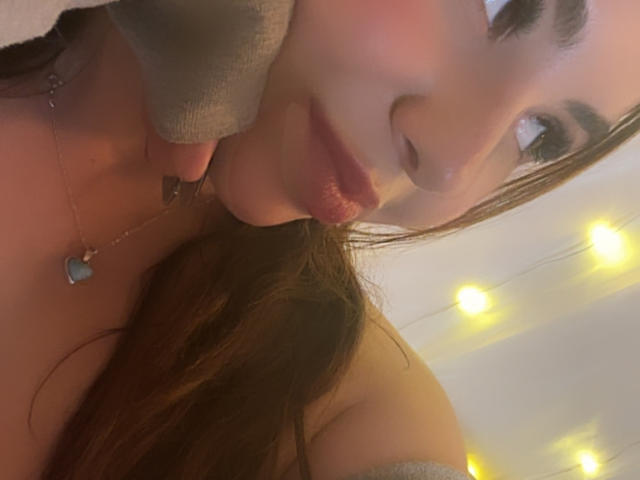 VenusxMusa with pouty lips, brown hair, and a grey off-shoulder top, wearing a heart necklace, and soft lighting in the background. Her face is slightly tilted.