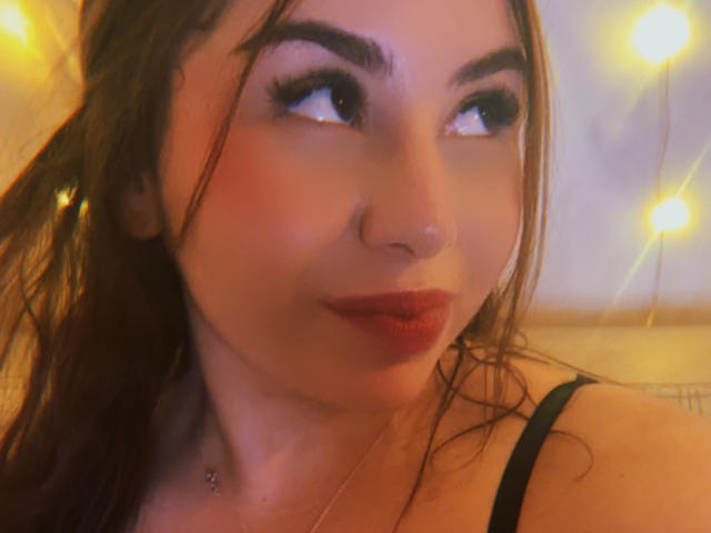 VenusxMusa mit rotem Lippenstift, dunklen Augenbrauen und langen braunen Haaren, die zur Seite schaut, trägt einen Nasenring und ein schwarzes Band, in weichem, gelbem Licht..