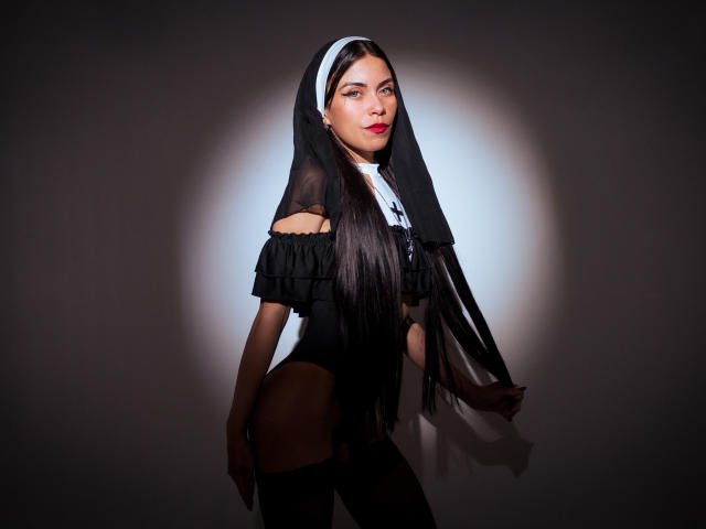 ZoeLux with long black hair, wearing a black nun outfit with a veil, off-shoulder top, and black thigh-highs, showing her tits and ass, standing against a bright light.