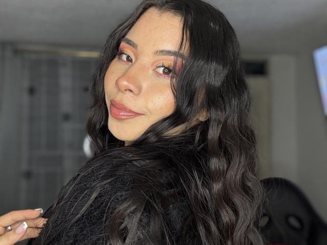 NatWatson has wavy black hair, pink eyeshadow, and pink lipstick. She's looking over her shoulder with a slight smile, wearing a black top. Her hand is near her face.