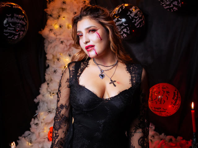 BiancaRey with bloody face, black lace dress, big boobs, and red lipstick, wearing a spider necklace, in a spooky Halloween setup with candles and decorative balls.