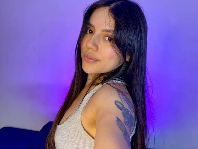 SophiaMarx with long black hair, gray tank top, tattoo on right arm, smiling at camera, purple-blue background, cute, natural makeup, medium tits, soft light.