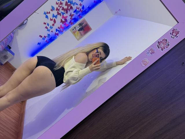 SammyViolet mit langen blonden Haaren, Brille, einem Pullover, der von einer Schulter fällt, schwarzen, hochgeschnittenen Unterhosen, macht ein Selfie vor einem Spiegel und zeigt ihr großes Gesäß und ihre dicken Oberschenkel, in einem Raum mit blauem LED-Licht..