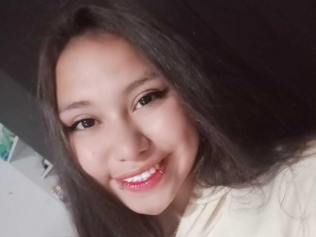 Annya Martinez tem cabelos longos e escuros, olhos castanhos e um sorriso grande com lábios vermelhos brilhantes. Ela está vestindo uma blusa branca, e seu rosto está centralizado na imagem..