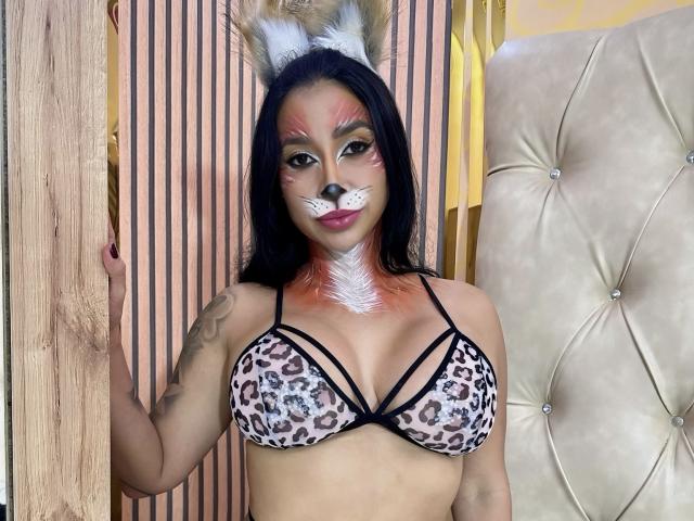FransheskaParker with leopard print bra, white furry face, rabbit ears, tattoo on arm, standing in front of wooden and tufted beige wall, black hair, orange-tinted hair tips.