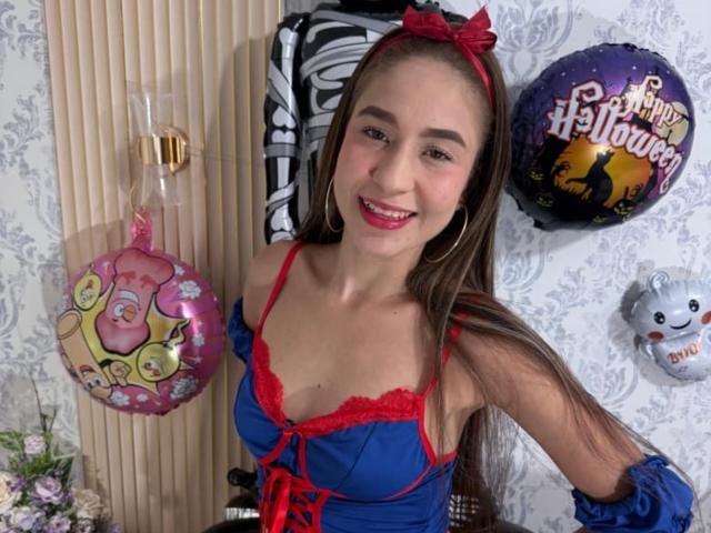 SammanthaOlssen, with long brown hair, wearing a red and blue Halloween costume, smiling, has small tits, and is standing next to Halloween balloons and decorations.