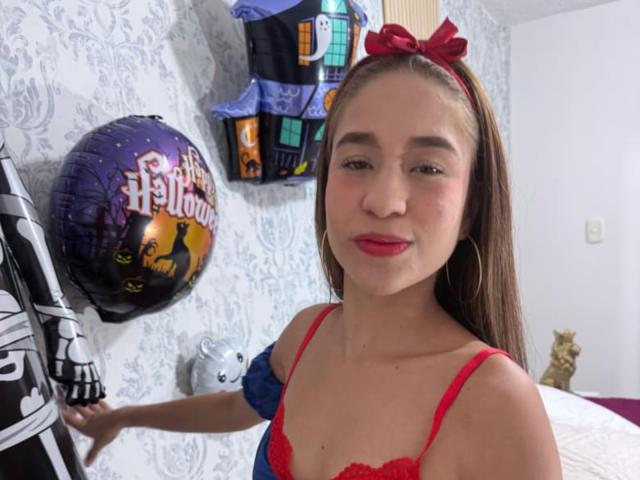 SammanthaOlssen with red lips, red bra, red bow, and long brown hair, holding Halloween balloons, wearing hoop earrings, smiling slightly, in a bright room.