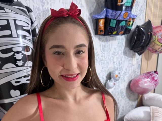 SammanthaOlssen with red hairband, hoop earrings, red lipstick, and red bra straps, smiling at the camera, showing her tits slightly in a cute, sexy way.