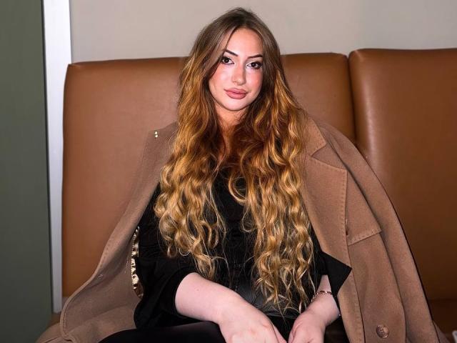 ElviraCrichlow with long wavy blonde hair, wearing a tan coat over a black dress, sitting on a brown leather couch, looking at the camera with a slight smile.