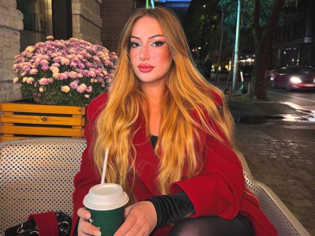 ElviraCrichlow with long blonde hair, red coat, and glasses, holding a coffee cup, sitting on a metal bench, pink flowers in a wooden box behind her.