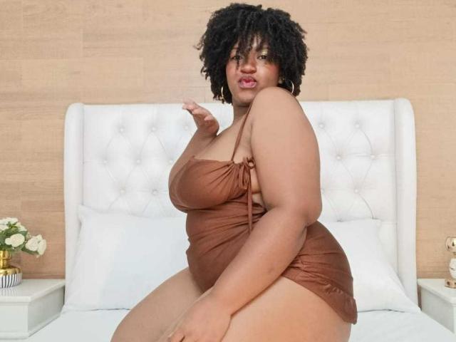 ViaraMoon, a curvy woman with dark curly hair, sits on a bed wearing a brown, strapless dress, showing her large, round ass and small, visible tits.