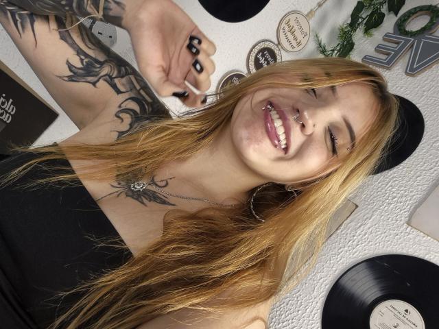 MailynnSkye with long blonde hair, black nail polish, nose ring, smiling, showing tattoos on arm and chest, wearing black top, black hat, and hoop earrings.