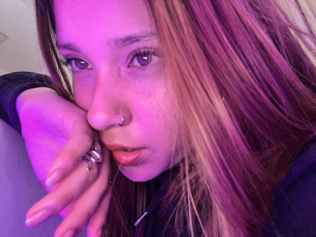 HeraJuno has pinkish-purple hair, a nose ring, and a ring on her hand, with a pensive expression, under purple lighting. Her lips are slightly parted.