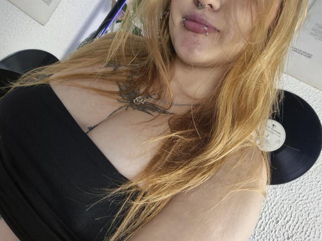 MailynnSkye with long, messy blonde hair, nose ring, lip piercing, and black tube top showing big tits and a tattoo below her collarbone. Vinyl records in the background.