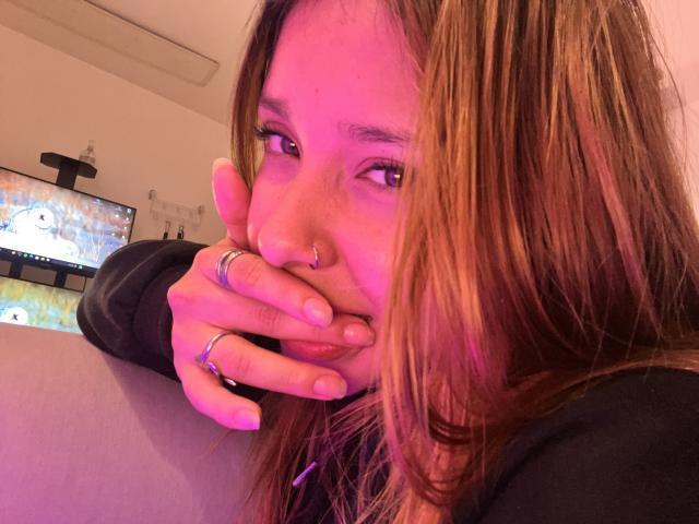 HeraJuno with brown hair, nose ring, and multiple rings, covering her mouth with her hand, in a purple-lit room, wearing a black top, looking slightly to the side.
