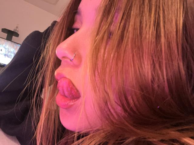 HeraJuno's face, pinkish light, brown hair, nose ring, tongue out, puckered lips, slight blush, black shirt, close-up, relaxed, casual, slightly messy hair, small nose piercing visible.