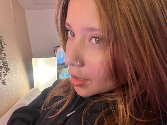 HeraJuno with brown hair, nose ring, and shiny lips, wearing a black hoodie, looking to the side with a slight smile. Soft light from the left.