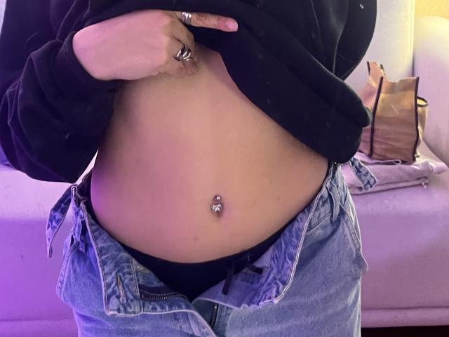 HeraJuno's belly button with a silver piercing, wearing unbuttoned blue jeans, black underwear, and a black sweater lifted to show her flat, tan-stained stomach.