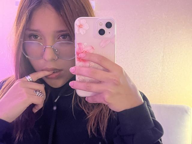 HeraJuno with long brown hair, glasses, nose ring, black hoodie, finger in mouth, holding phone with pink flower case, looking at mirror, pink and purple lighting.