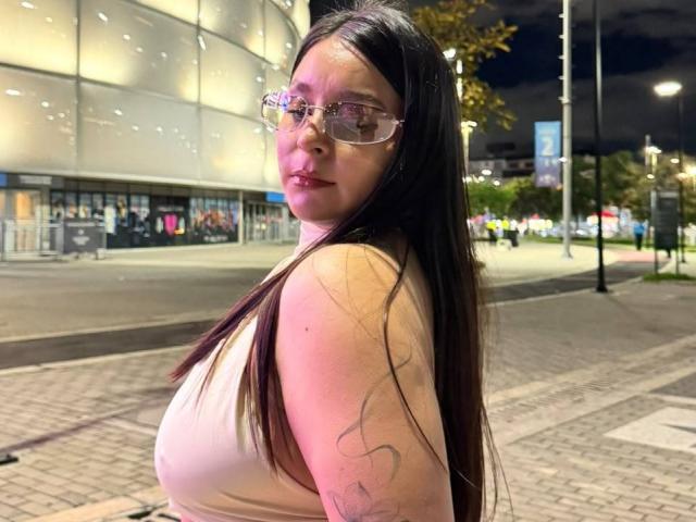 EmaJames with long black hair, glasses, and a pinkish tattoo on her right arm, wearing a beige tank top, standing on a nighttime city sidewalk.
