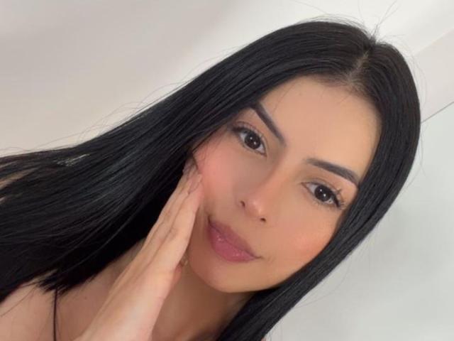IsabellaJames has long black hair, brown eyes, and is touching her cheek with her hand. She's smiling with pink lips. Her eyebrows are thick and well-groomed.
