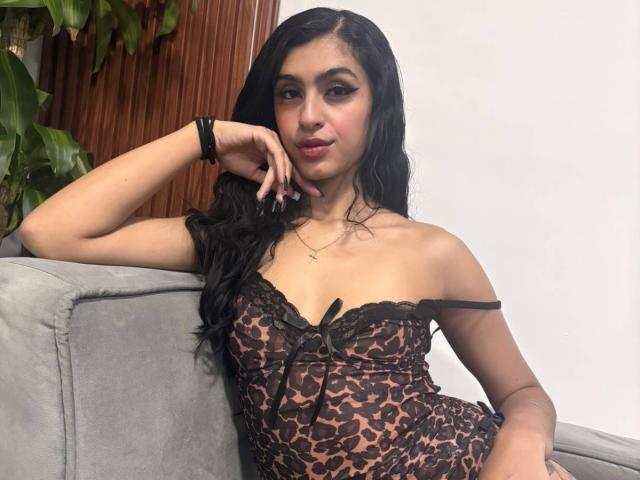 LilithDesire has long black hair, wearing a leopard-print dress with black lace, showing her small tits, sitting on a gray couch, arm resting on it, looking at camera.