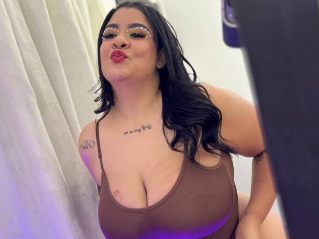 RashaelBig with big tits, wearing a brown tank top, black hair, pouty lips, and sparkly eye makeup, leaning forward, showing tattoos on chest and arm.