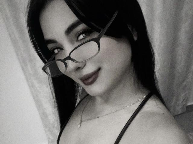 Zwart-wit selfie van Stephaniy69 met een bril, donkere lipstick, lang zwart haar, een ketting, lachend, waarbij haar borsten zichtbaar zijn in een top met spaghettibandjes..