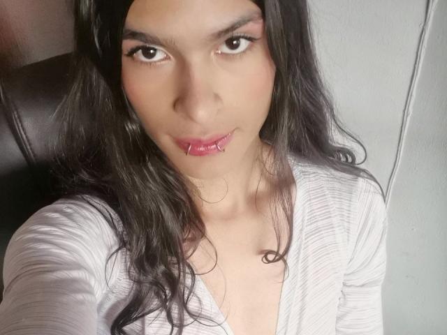 LunarisSmith has long black hair, brown eyes, and pink glossy lips, wearing a white ribbed top with a slight V-neck, sitting on a dark leather chair.