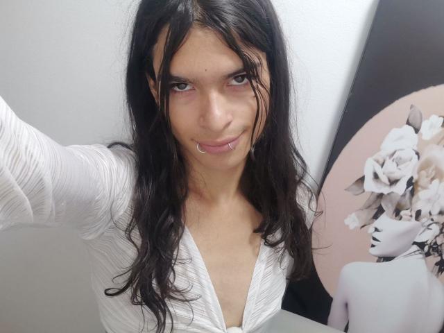 LunarisSmith with long black hair, wearing a white V-neck shirt, showing a nose ring and smiling at the camera, has a medium build and a cute face.
