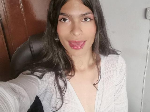 LunarisSmith has dark hair, brown eyes, and pink lipstick with her tongue out, wearing a white, deep V-neck top, showing a bit of cleavage, sitting in a black chair.