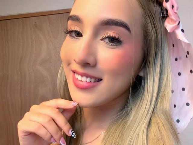 EmmaWat with blonde hair, pink polka-dot bow, smiling, pink lipstick, rosy cheeks, long eyelashes, finger with white nail and black skull design, holding her chin.