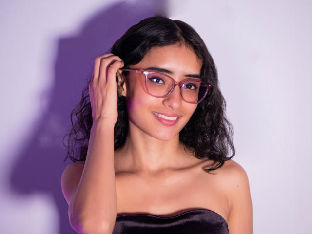 Layla Brooks, con cabello rizado oscuro, piel morena y gafas rosas, sonriendo mientras se toca la oreja, vestida con un top negro sin tirantes, mostrando sus pequeños senos y su cara linda..