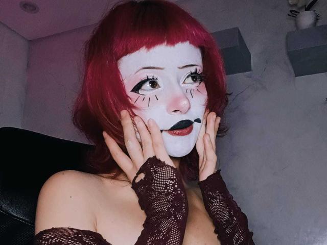 ViolettClay with red hair, white face paint, black lipstick, and lace sleeves, hands on cheeks, looking up with big eyes, pale skin, and visible cleavage in a dim room.