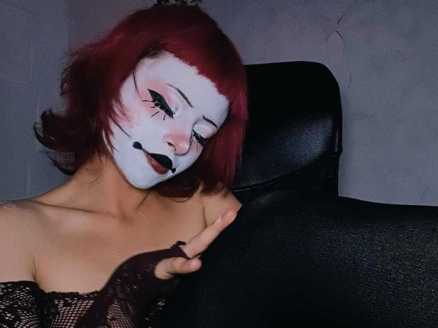 ViolettClay with white face paint, red hair, black lipstick, and eye makeup, pointing with a finger, wearing a black mesh top, in a black chair.