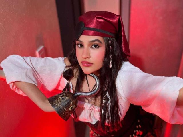 IrisBlair with long dark hair, wearing a red satin hat, white puffed sleeves, black corset, and brown metal glove, holding a pirate hook, red lighting.