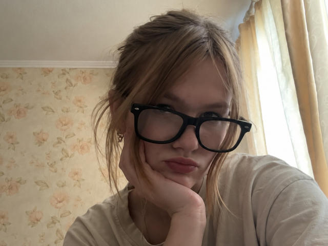 SeeOlausen, avec ses cheveux blonds en bataille, ses lunettes noires et ses lèvres fines, le menton appuyé sur sa main, portant une chemise beige, a l'air légèrement agacé ou ennuyé..