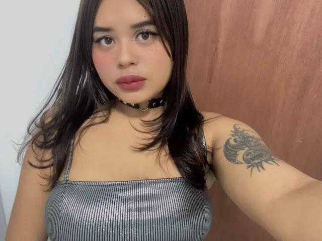 ChiaraBella with dark brown hair, wearing a silver ribbed tank top, black choker, and pink lipstick, showing a dragon tattoo on her right arm. Her tits are small.