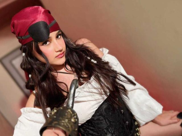 IrisBlair with long dark hair, red hat, eye patch, white off-shoulder shirt, black corset, holding a brass bell, looking up with a slightly pouty expression.