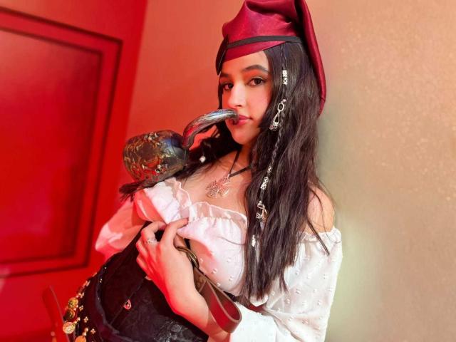 IrisBlair with long black hair, wearing a red beret, white off-shoulder top, black corset, holding a hookah, sucking on a pipe, looking at camera, indoors, red lighting.