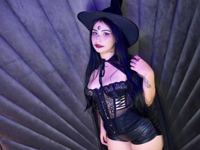 HannahBecker with long black hair, witch hat, dark lipstick, black lace corset, and leather shorts, standing with a hand on her hat, showing tattoos and a cute face.