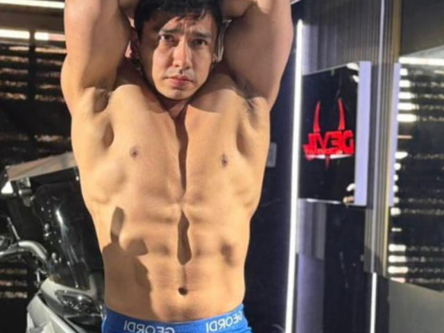 EithanConor, musclé avec des abdominaux bien dessinés, portant un sous-vêtement bleu, les bras levés, debout dans une salle de sport, avec une expression sérieuse. Sa peau bronzée et ses cheveux courts et foncés sont remarquables..