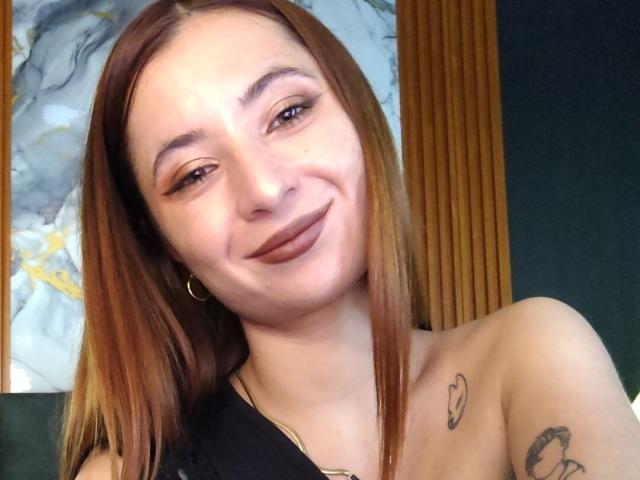 Lizzie Allen mit hellbraunen Haaren, die lächelnd mit dunklem Lippenstift, kleinen Reifenohrringen, Tattoos an den Schultern und nackten Schultern in die Kamera schaut..