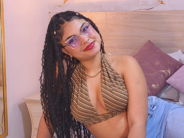 MelodyAlvarez with long black braids, wearing glasses, a brown striped halter top showing her tits, and blue jeans, smiling at the camera. She has red lipstick.
