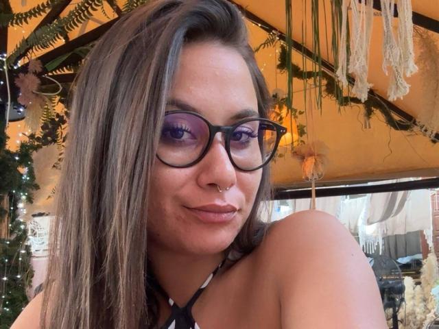 JessieBoo has brown hair, purple eyeshadow, black glasses, a nose ring, and is pouting with her topless shoulders visible. Her tits are not shown. She's in a cozy, plant-filled room.