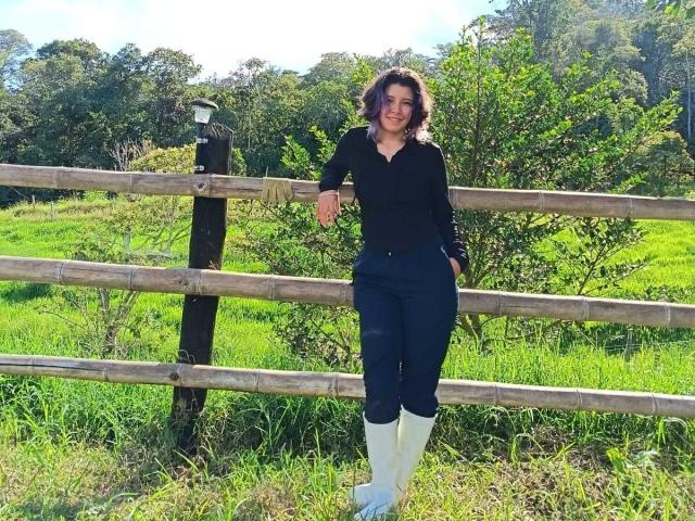 DarckVelvet, standing with arms relaxed on a wooden fence, wearing a black long-sleeve shirt, navy pants, and white knee boots, smiling with curly brown hair, in a sunny green field.