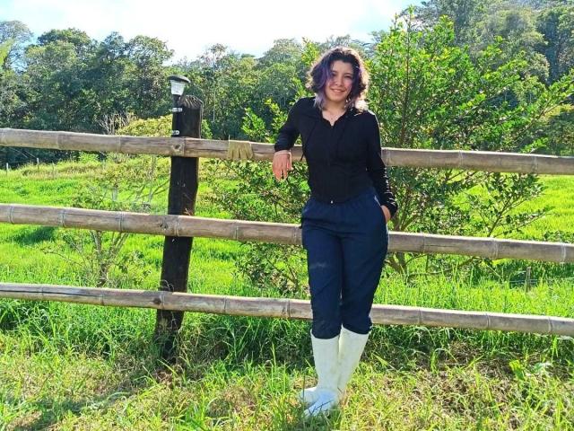 DarckVelvet stands with one arm on a wooden fence, wearing a black shirt, navy pants, and white boots, smiling with curly purple-tinted hair, in a sunny green field.