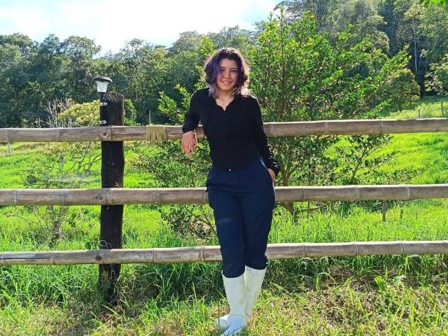 DarckVelvet stands in front of a wooden fence, wearing a black top, blue pants, and white boots, with a hand in her pocket, smiling. Green grass and trees behind.